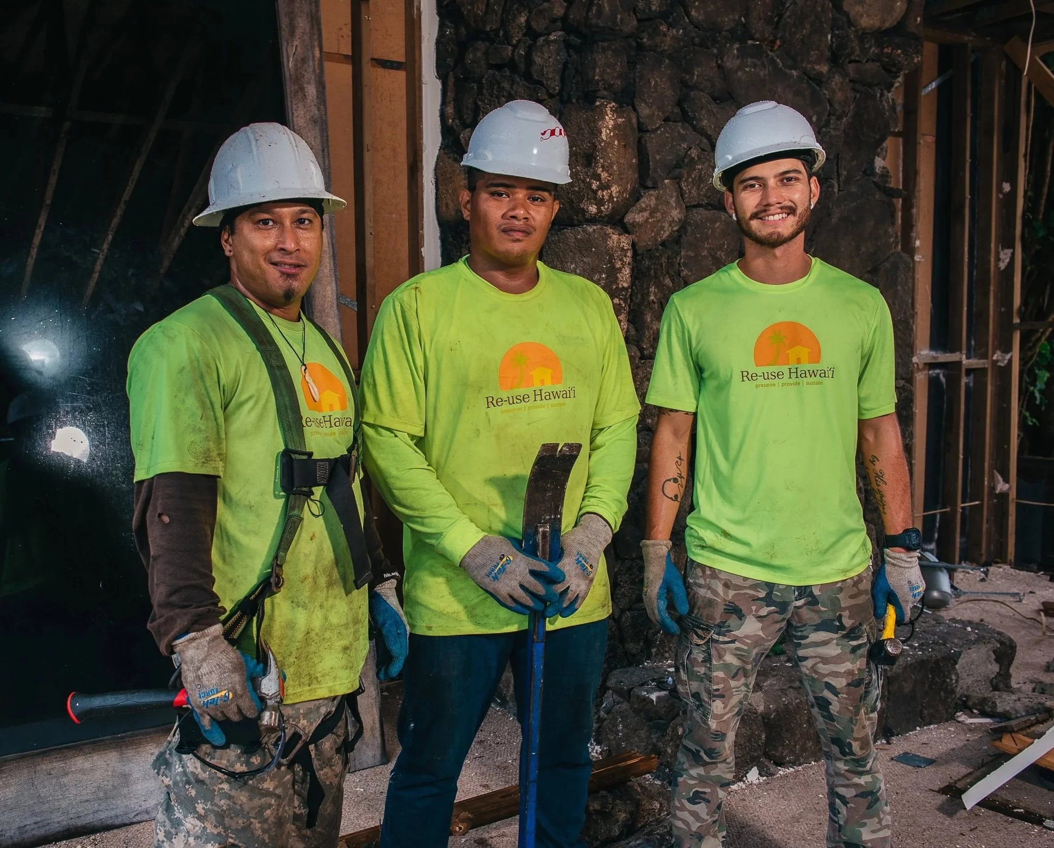 Three men wearing construction helmets and bright yellow shirts, with two of them also in camouflage pants, standing in front of a stone wall and building framework, smiling and holding tools.