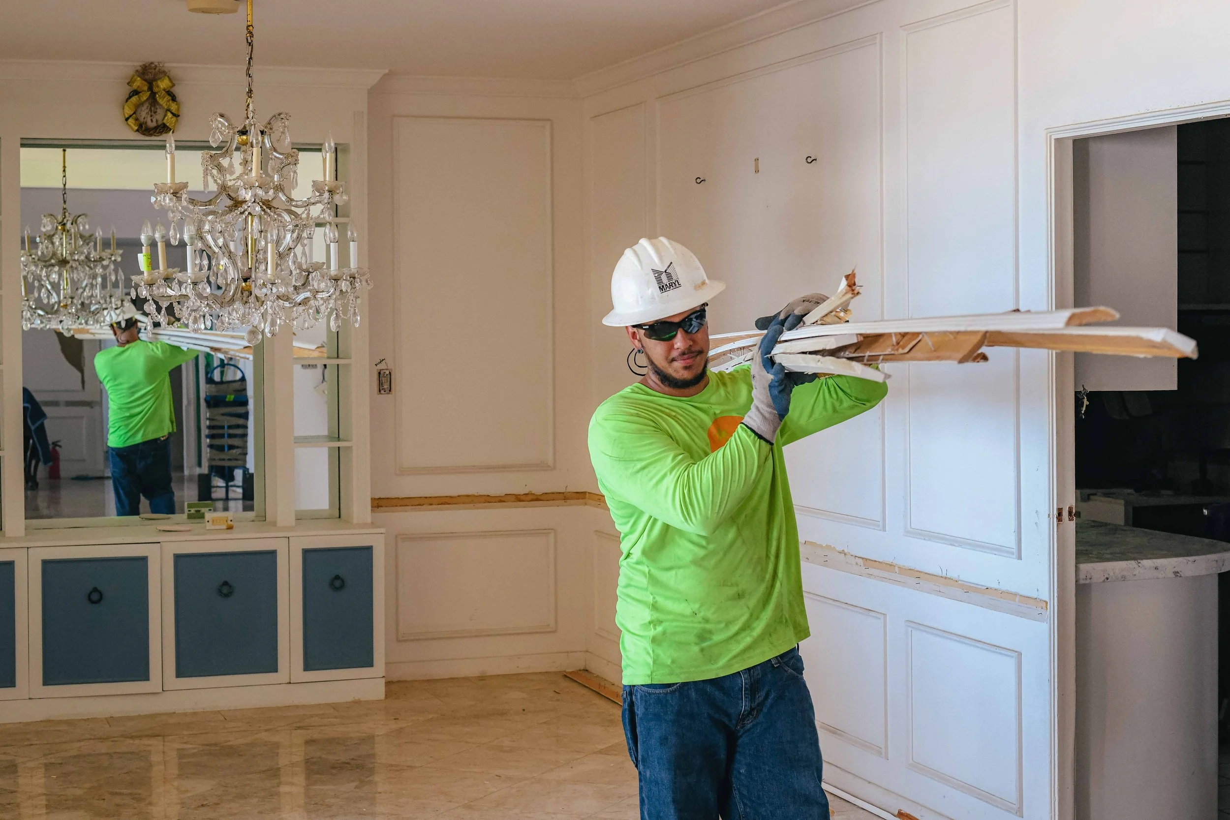 A man carrying a stack of long lumber on his shoulders in a home that is being remodeled.
