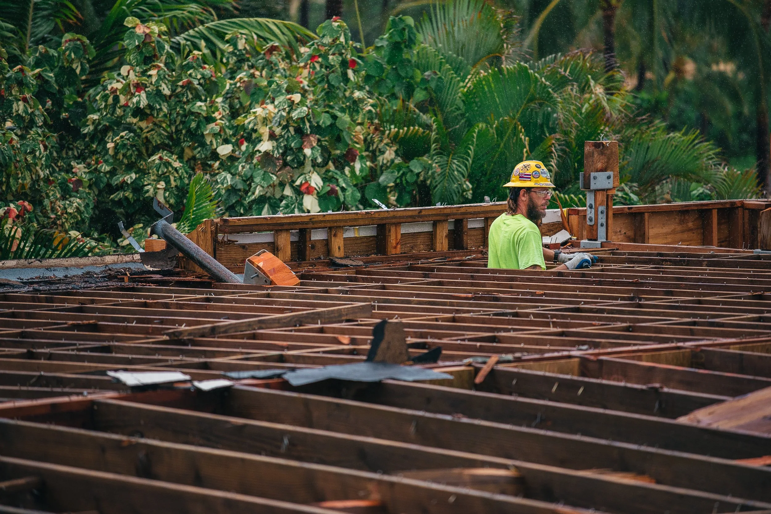 Construction worker wearing a yellow helmet and green shirt working on a wooden structure, with lush green tropical plants in the background.
