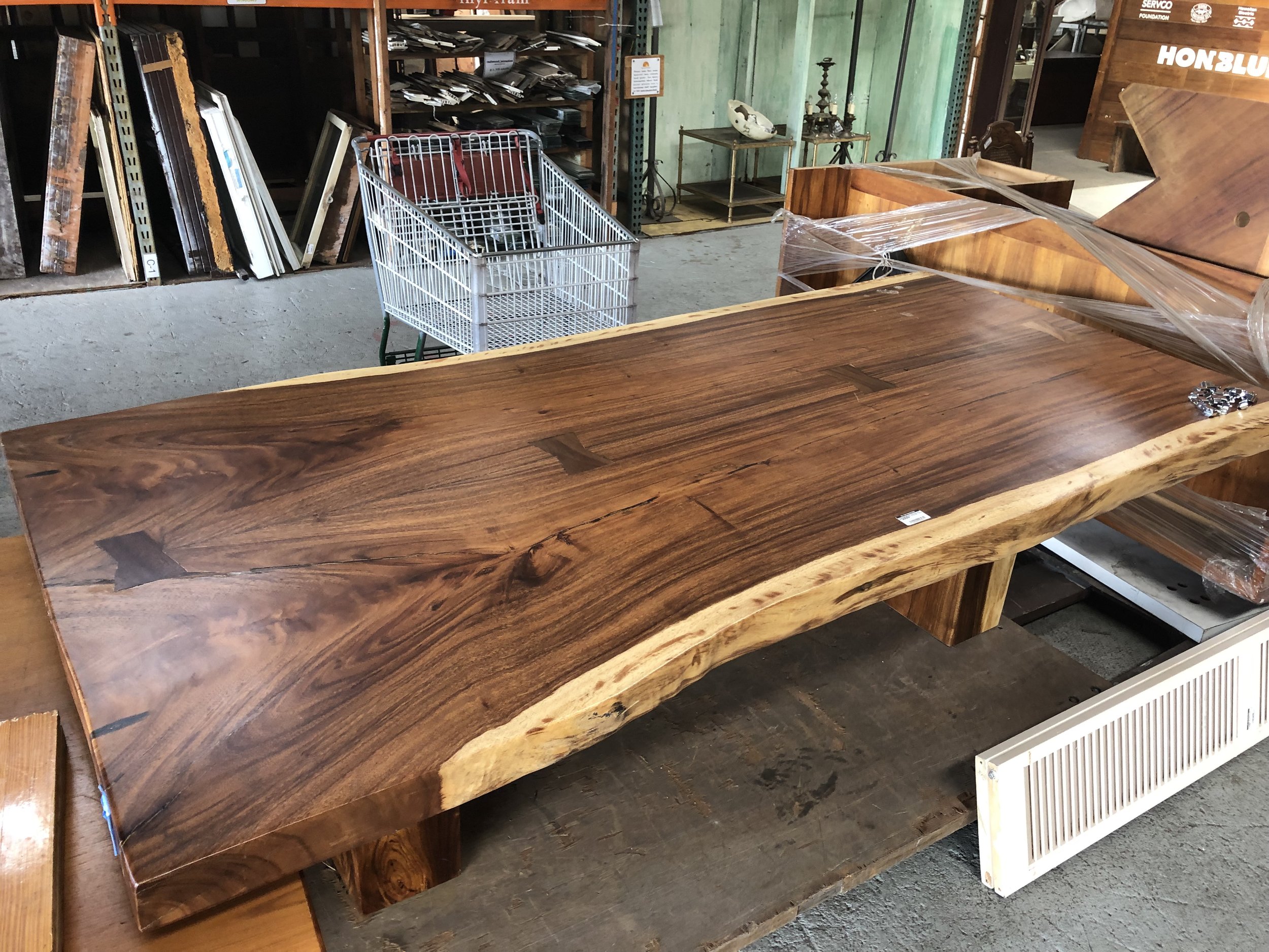 A large, polished wood table with natural, uneven edges sits in a warehouse or store. Behind the table, there is a shopping cart and shelves with various other furniture pieces and materials.