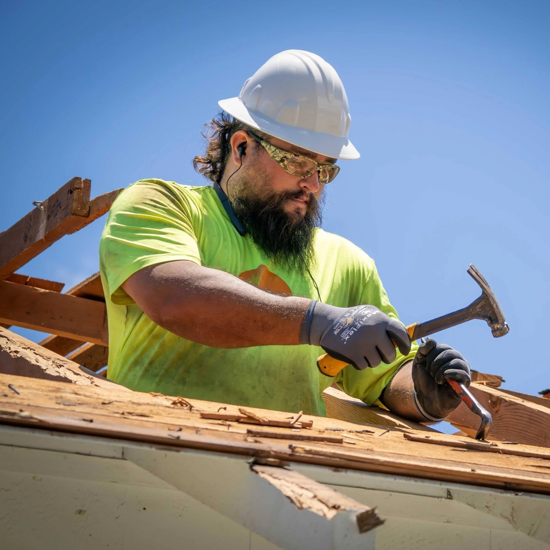 A construction worker wearing a white hard hat, yellow shirt, gloves, and safety glasses is hammering nails into a wooden structure on a sunny day.