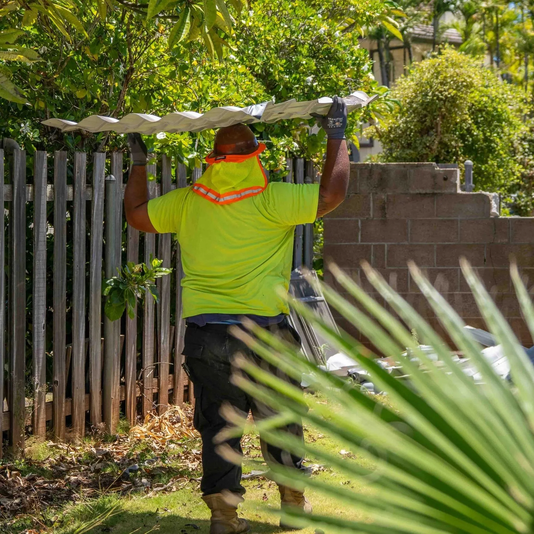 A worker wearing a yellow safety vest, black gloves, and a wide-brimmed hat is holding a long, curved metal blade above his head in a backyard with a wooden fence, brick wall, and green trees.
