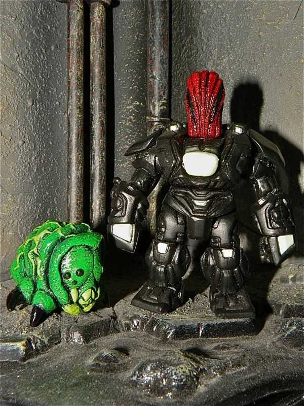 A small green artificial creature resembling a frog or bug on the left and a larger black robot figure with red hair on the right, both placed on a rocky surface in front of a dark textured wall with pipes.