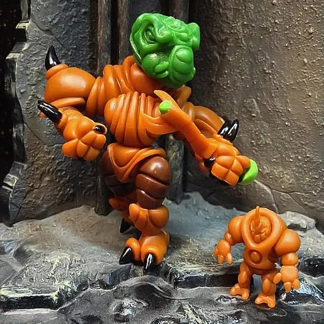 Colorful clay or plastic figurine of a monster with green hair, orange body parts, black claws, and brown accents, standing on rocky terrain.