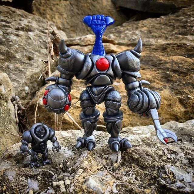 Two toy robots with a rocky outdoor background. The larger robot is dark gray with a red torso and a blue arm and head. The smaller robot is also dark gray, with red details, and has a rounded body.