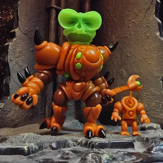 Colorful toy figurines resembling monsters with horns and claws, including a larger one with a green skull and a smaller one holding a staff, standing on a rocky surface with a textured background.