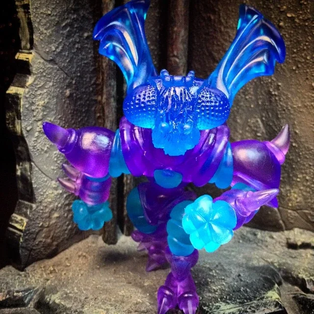 Colorful, translucent sculpture of a fantastical creature with a dragon-like face, large wings, multiple limbs, and floral details, illuminated in blue and purple hues.