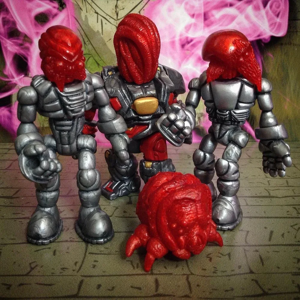 Action figures resembling robots with red alien-like heads, one in the center with a suit and two on the sides with silver armor, standing on a wooden surface with a pink and purple smoky background.