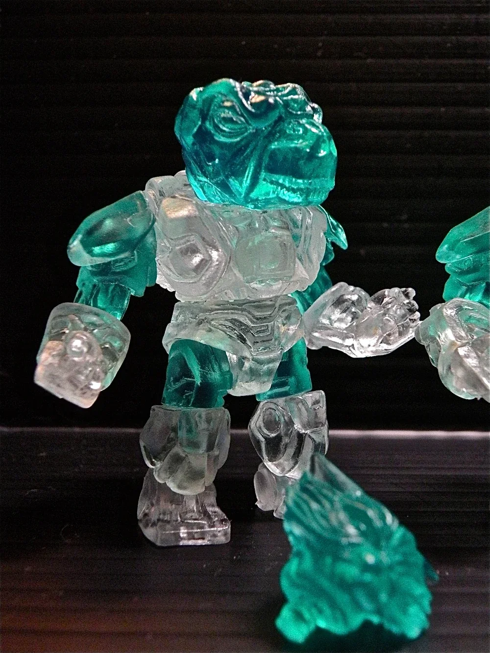 A plastic toy figure resembling a robot or monster with transparent blue and clear parts, standing on a black surface with a textured black background.