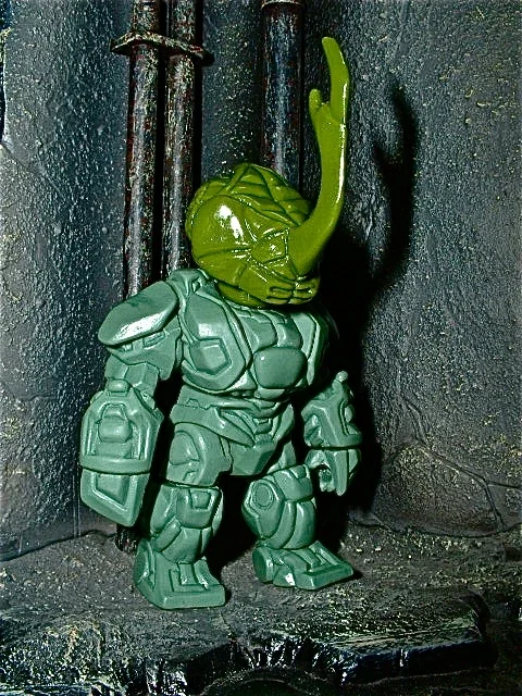 A toy figure combining a green humanoid with plant-like features and armor, standing against a dark textured wall and rusty pipes.