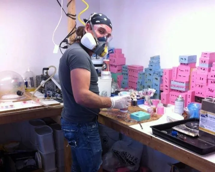 Person wearing a mask, goggles, and gloves working at a cluttered workshop table filled with colored boxes, tools, and supplies.