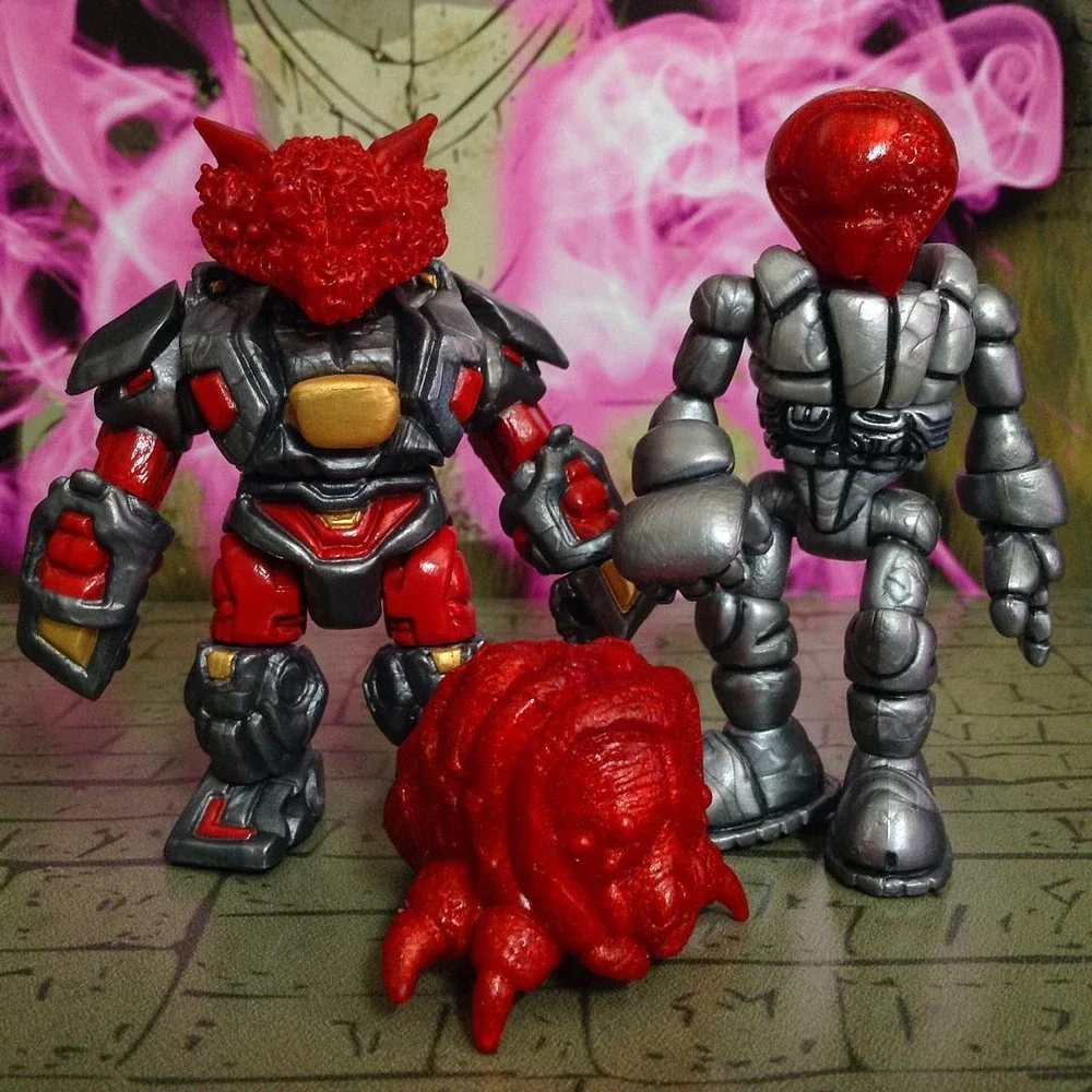 Two toy figures with unusual features. The figure on the left has a red, textured, animal-like head with horns, armor, and red and black clothing. The figure on the right has a shiny, red, heart-shaped head with a silver, robotic body. Between them o