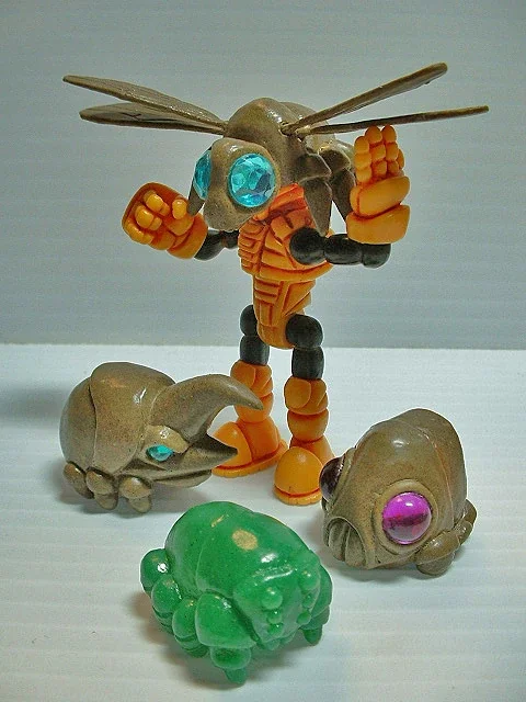 Toy robot with insect features and three small animal figures, including a green creature, a beige animal with a purple eye, and another beige animal with a blue eye, on a white surface.