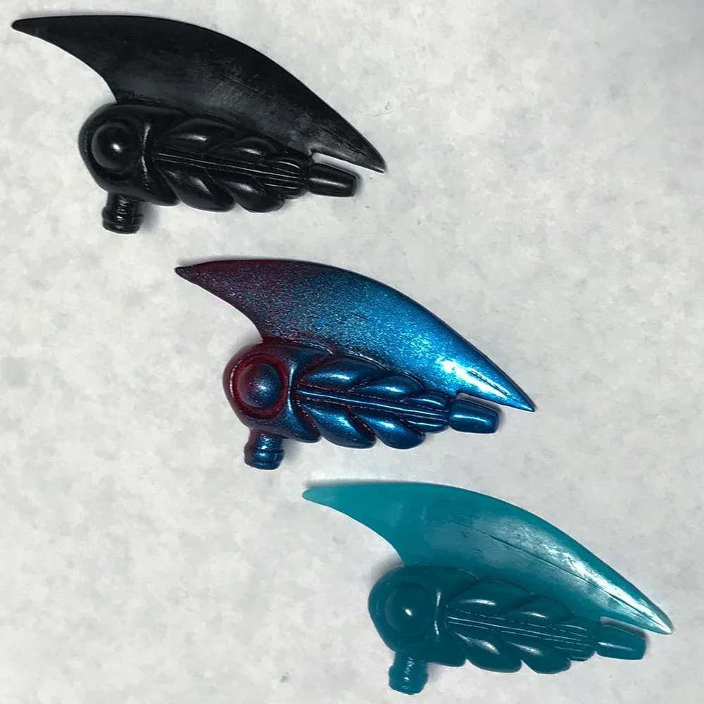 Three small, colorful, wing-shaped objects with a bead-like detail at their bases, arranged on a white surface.