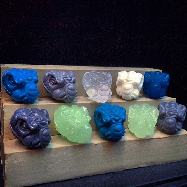 Assorted colorful soap bars shaped like tribal masks, arranged on a wooden display stand.