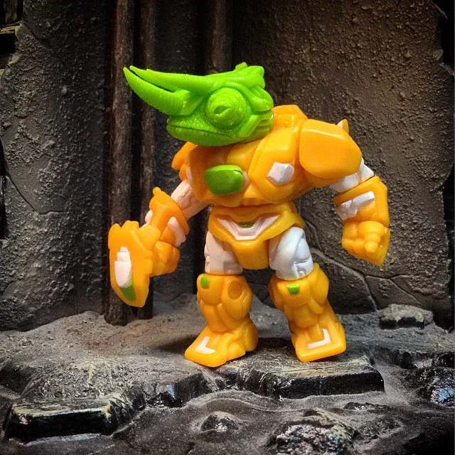 A toy robot with a green dragon head, orange and white armor, and yellow boots, standing on a textured surface against a dark background.