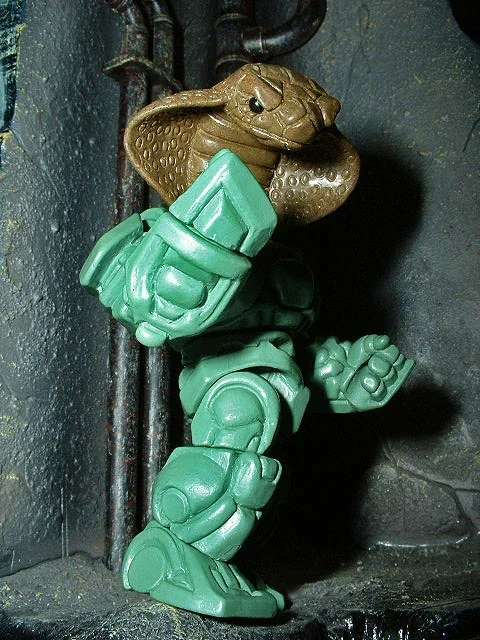 Stacked green and brown plastic toys resembling a robot and a dinosaur on a dark surface with pipes in the background.