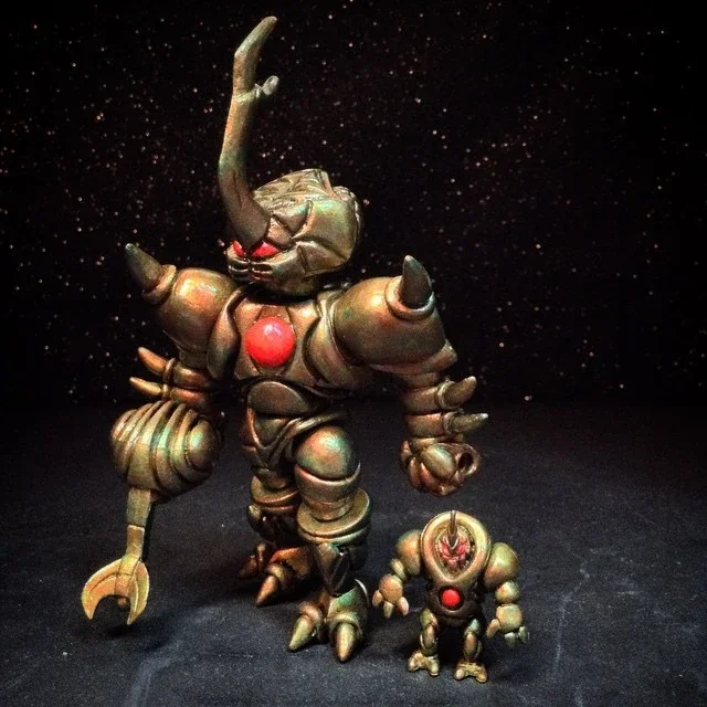 Two metallic robot figurines, one larger and one smaller, set against a black background with star-like specks. The larger robot has a horn on its head and appears to be reaching upward, while the smaller robot stands nearby.