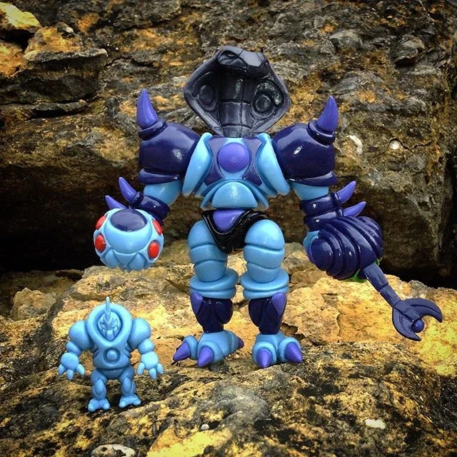Two blue robot action figures, one large and one small, posed on rocks against a rocky background.