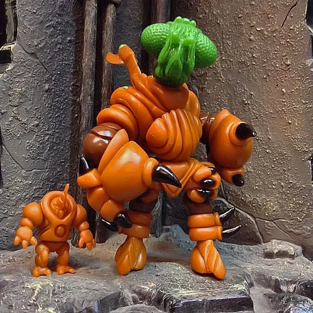 Orange and green toy figures resembling robots or monsters on a rocky surface against a textured wall.