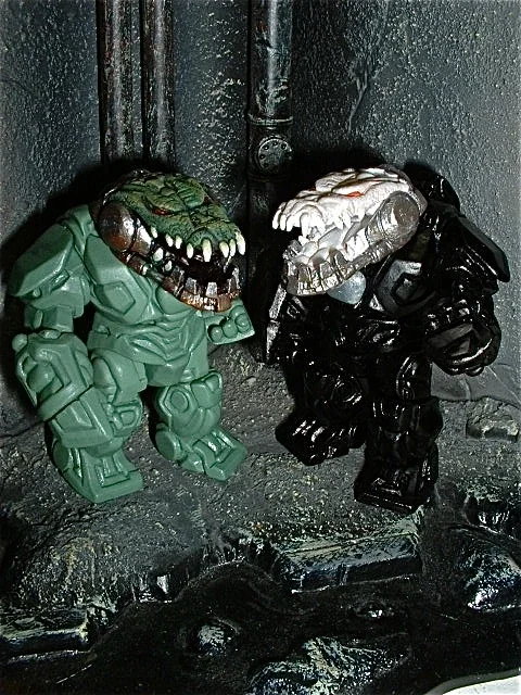 Two dinosaur-themed toy figures, one green and one black, with detailed skull heads, are positioned against a dark, textured background, possibly on a street or industrial setting.