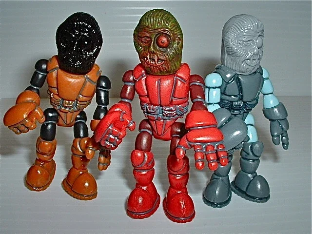 Three vintage robot action figures with monster heads, one orange, one red, and one gray, standing on a surface.