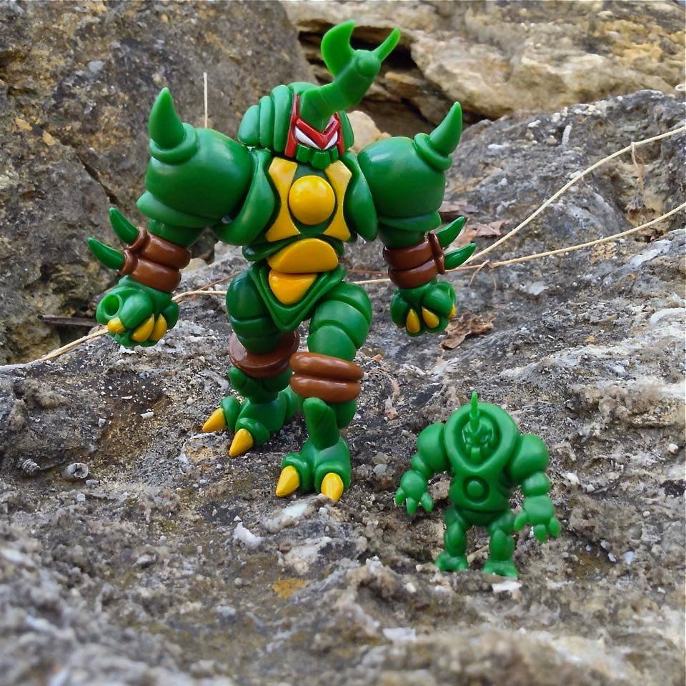 Two green toy monster figures on rocky ground, one larger with horns and spikes, the other smaller with a smooth head.