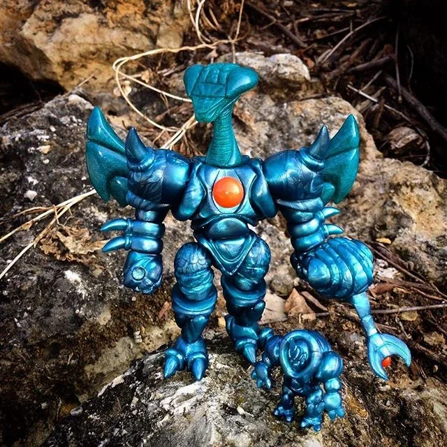 A blue action figure or toy robot with orange accents, standing on rocks outdoors.