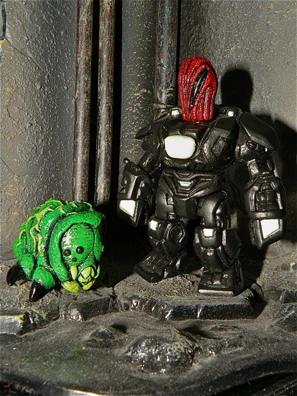 Two small toy figures: a green caterpillar and a black robot soldier with a red head, standing on a textured surface against a dark background.