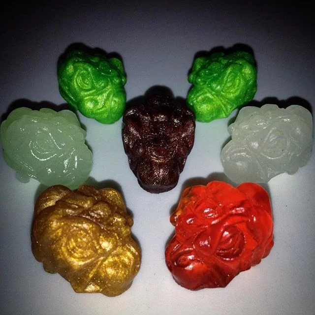 Six gummy candies shaped like faces, each in different colors: green, white, black, gold, red, and green. They are arranged in a semi-circle on a white surface.