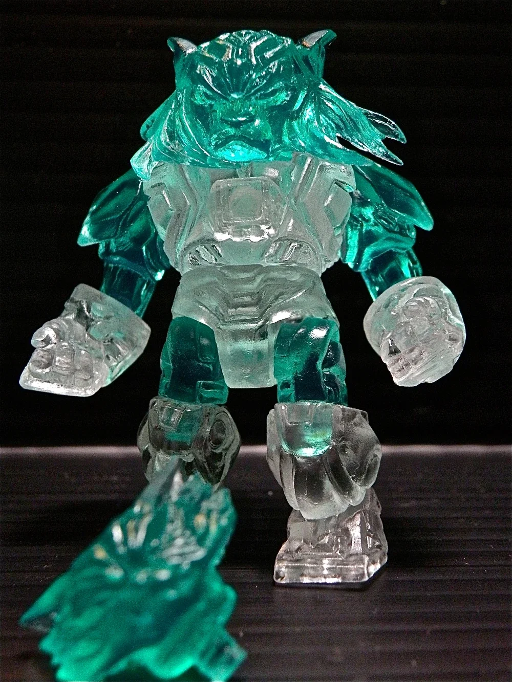 A glass figurine of a muscular humanoid creature with a lion-like face, standing on a black surface against a black background. The figure has translucent teal and clear glass parts, with a piece of teal glass sculpture in the foreground.