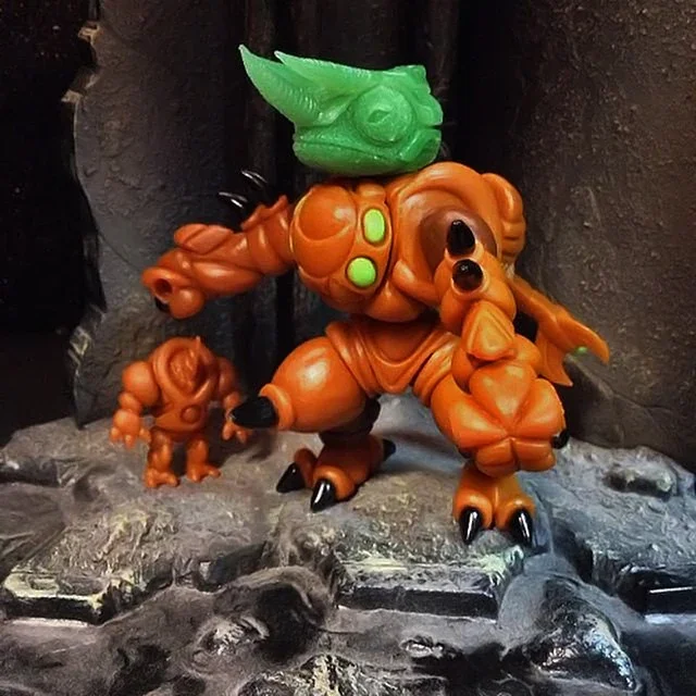 A toy figure resembling a large orange monster with green and black accents, holding a smaller orange figure, with a green headpiece, standing on a rocky surface.