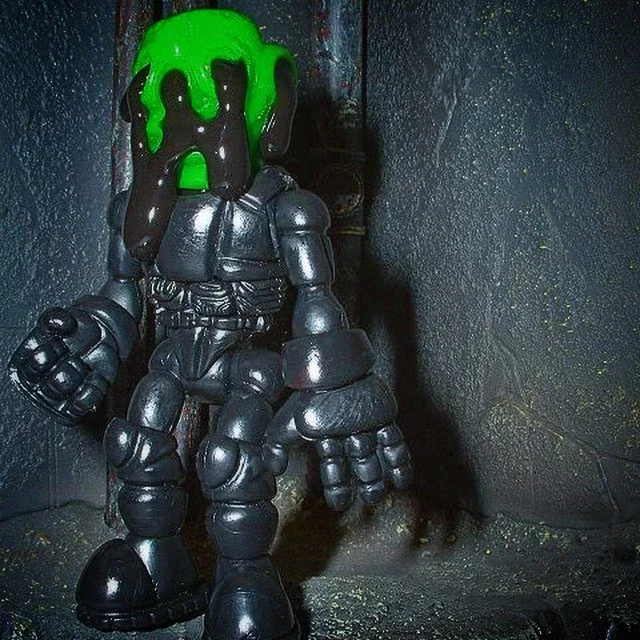 A chrome-colored toy robot with a green dripping substance on its head, sitting against a dark textured wall.