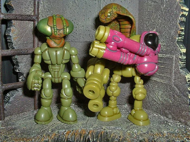 Two toy robot figures, one green and the other pink with a yellow head, standing on a brick surface against a wall.