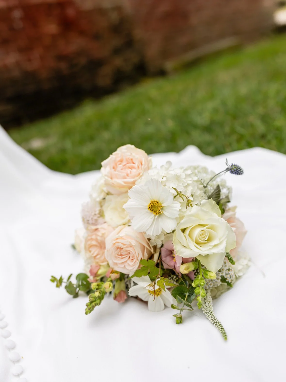 A bouquet of light pink, white, and cream roses, accented with white daisies, lavender, and green foliage, resting on a white fabric surface with a brick wall and grass in the background.