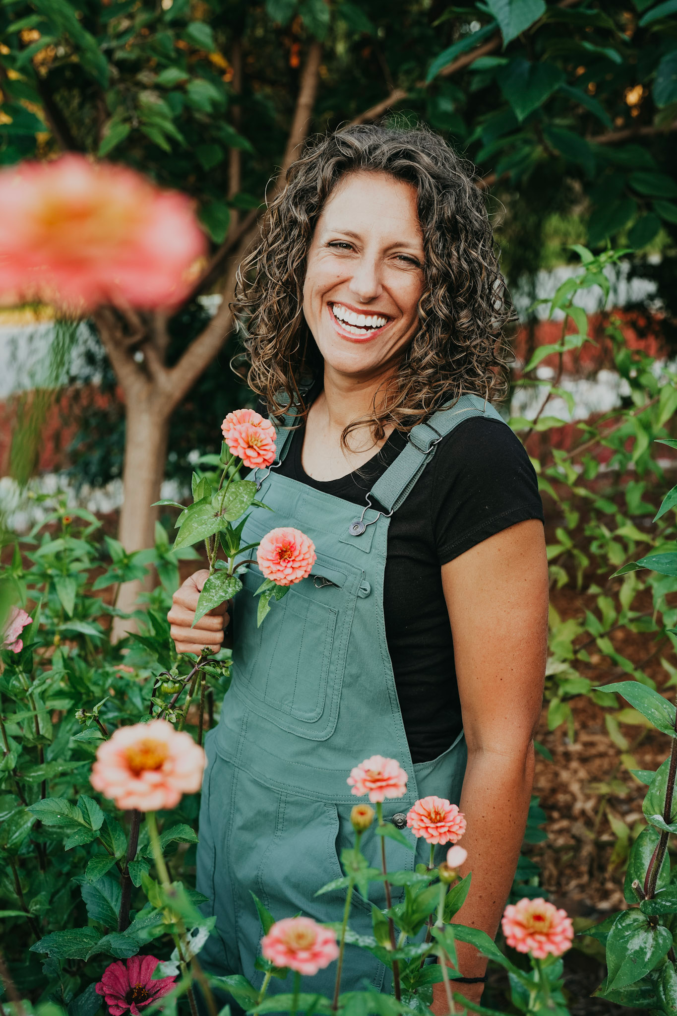 Woman with curly hair and a black shirt smiling, holding pink flowers in a garden.