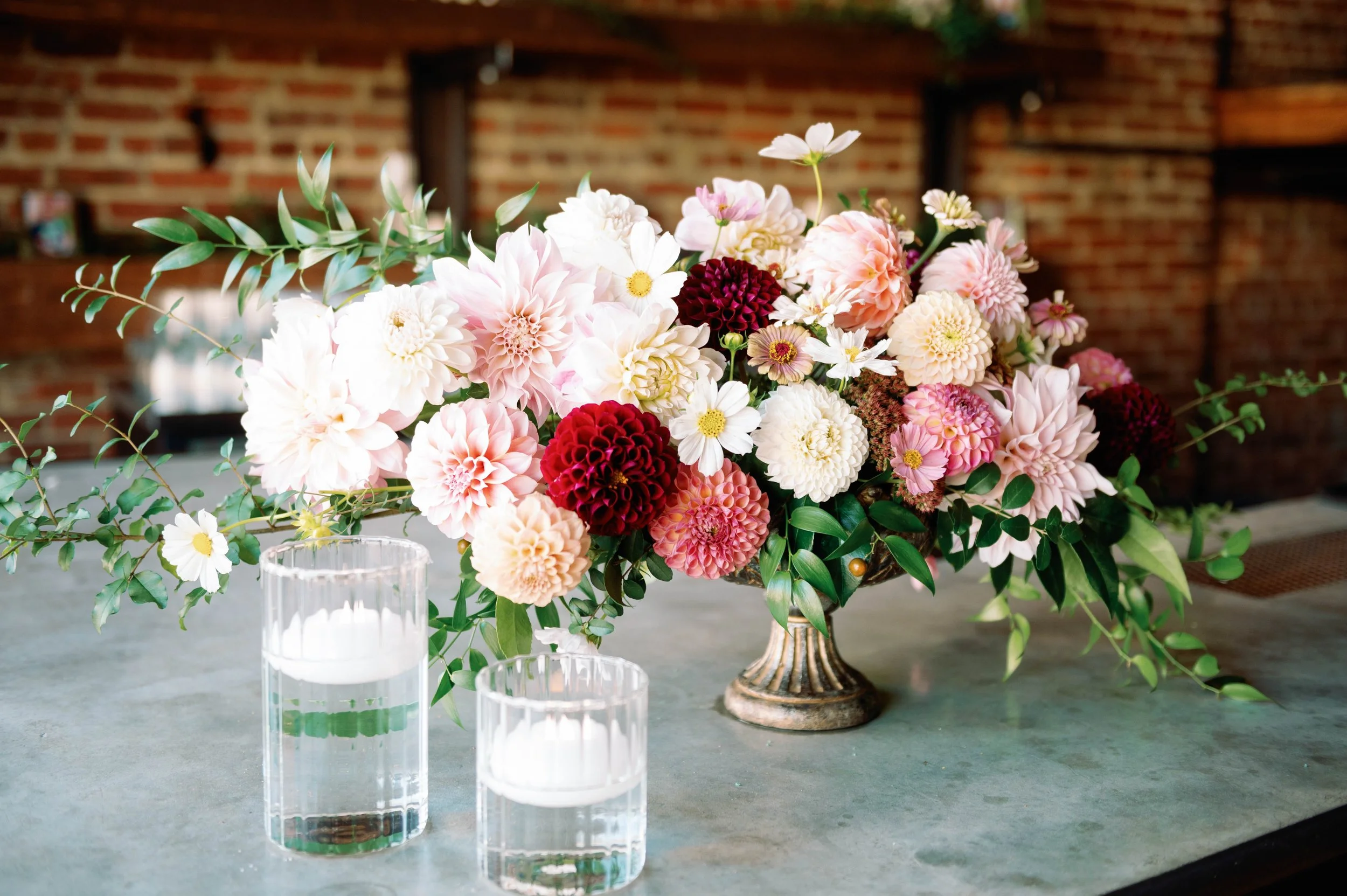 A large floral arrangement with pink, white, and deep red flowers in a decorative vase on a table, accompanied by two glass candle holders with white candles, set against a brick wall background.
