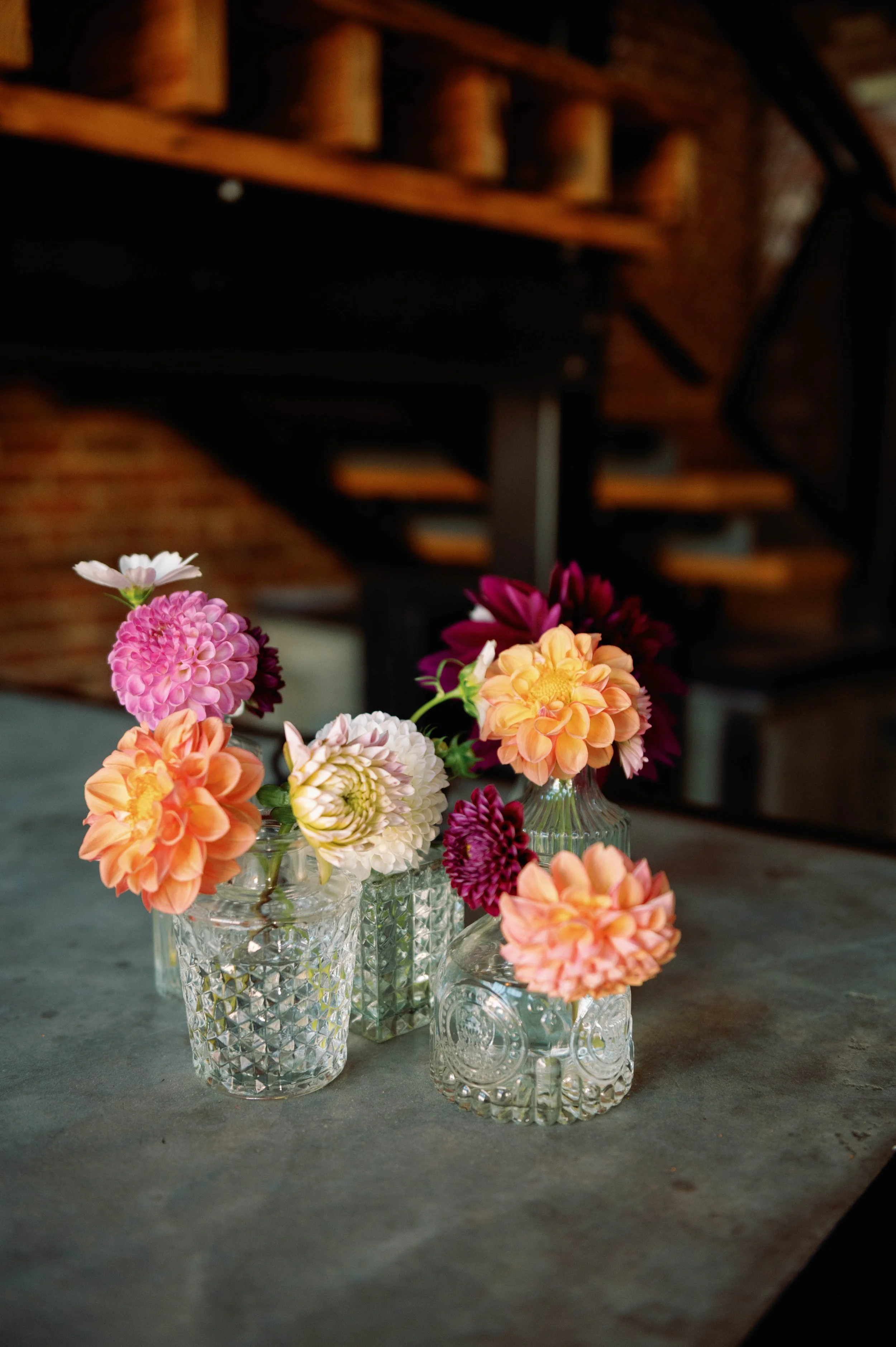 Several colorful dahlias in small glass vases on a concrete surface, with a blurry background of a brick wall and a wooden staircase.