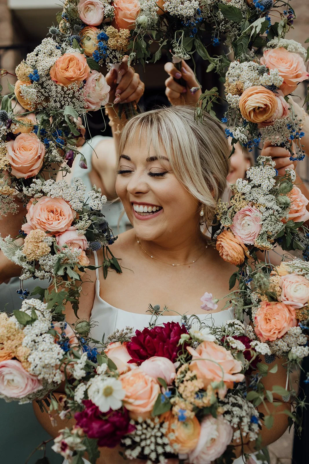 A smiling woman with blonde hair surrounded by a floral wreath and bouquet, holding it up with multiple hands.