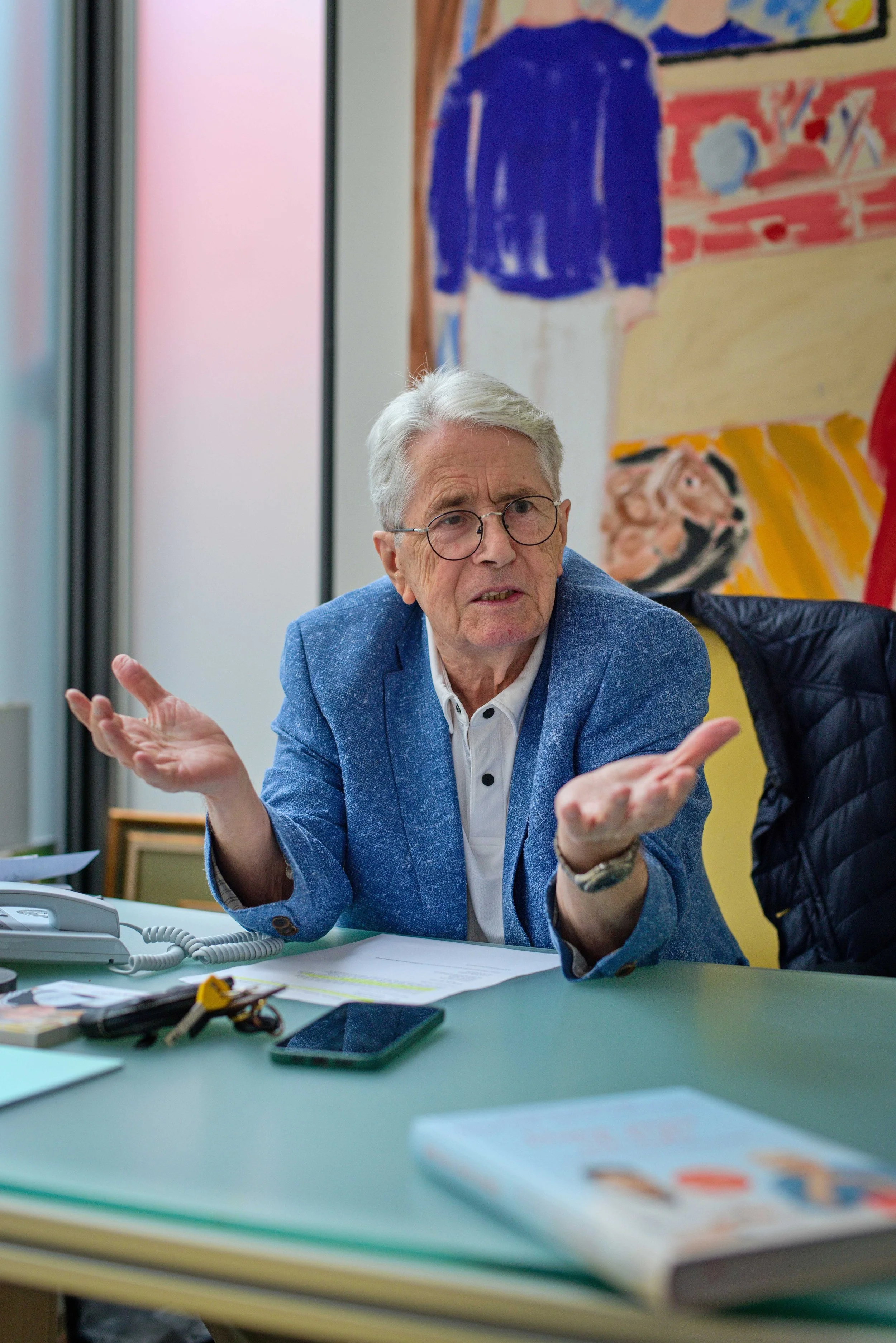 Frank Elstner sitzt an einem Tisch in einem Büro, trägt einen blauen Blazer und erklärt etwas mit einer ausladenden Handbewegung. Im Hintergrund hängen bunte Gemälde an der Wand. Frank Elstner während eines Gesprächs bei einer Veranstaltung.