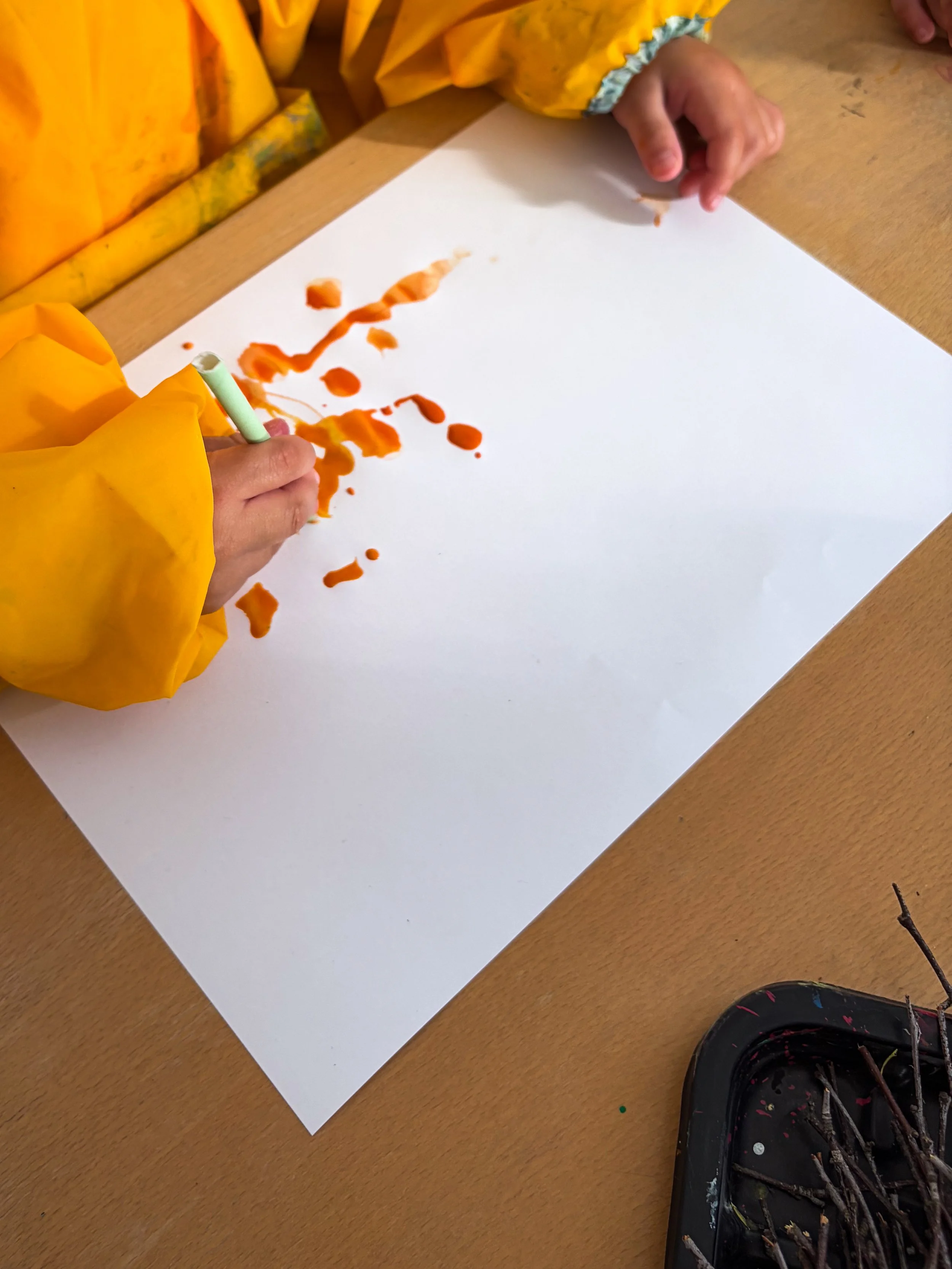 Barn i gula arbetskläder ritar på vitt papper med orange färg. Det finns ett svart plastlock med penslar och trästickor.