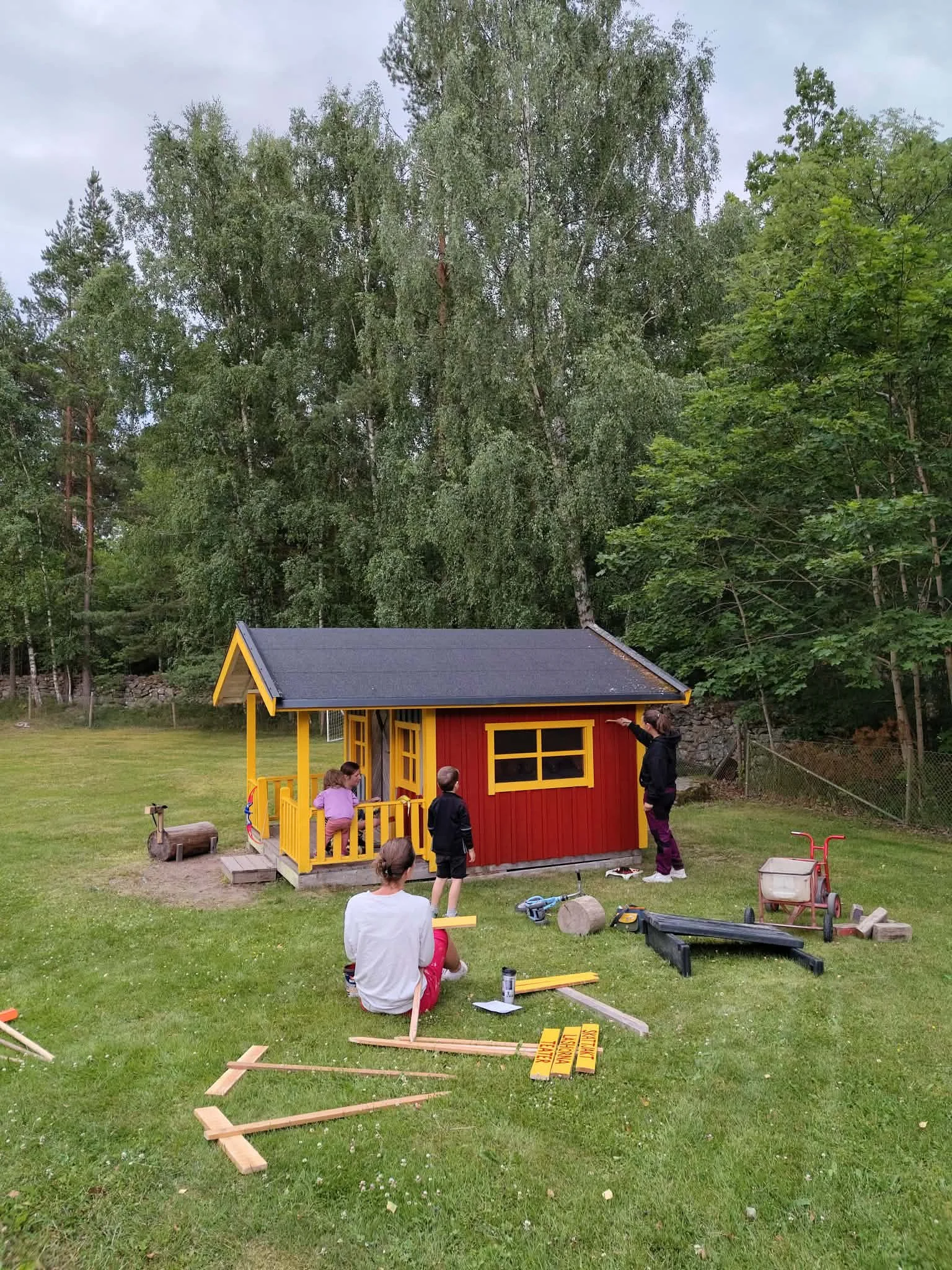 En liten lekstuga med röd vägg och guldkant, byggs av ett grupp människor utomhus. Flera personer, inklusive barn och vuxna, samlar material och arbetar på lekstugan. Plast, trä och byggverktyg ligger på gräset.