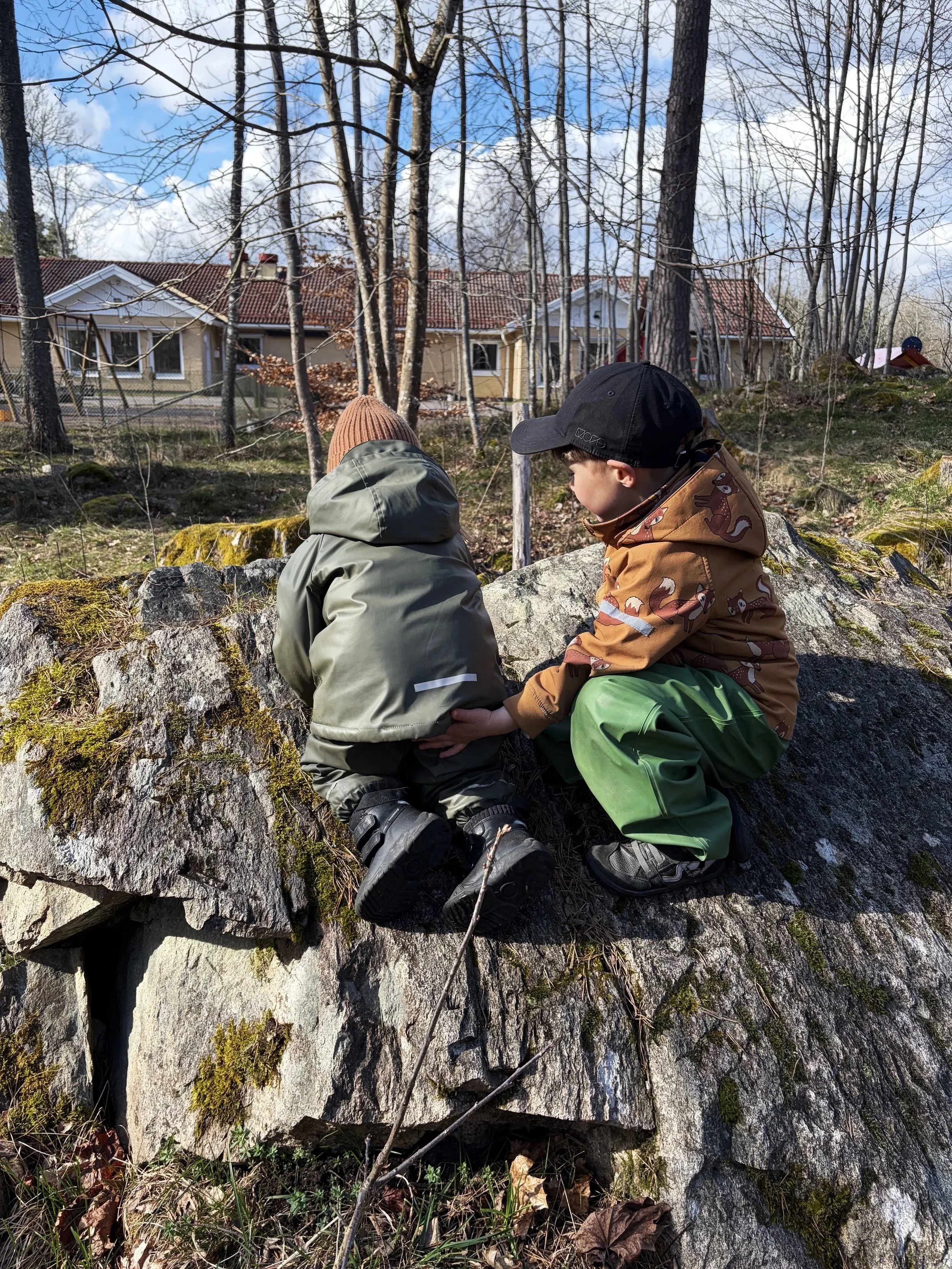 Två barn som sitter på en stor sten i en skog med träd och äldre villor i bakgrunden, båda har mössa och jacka.