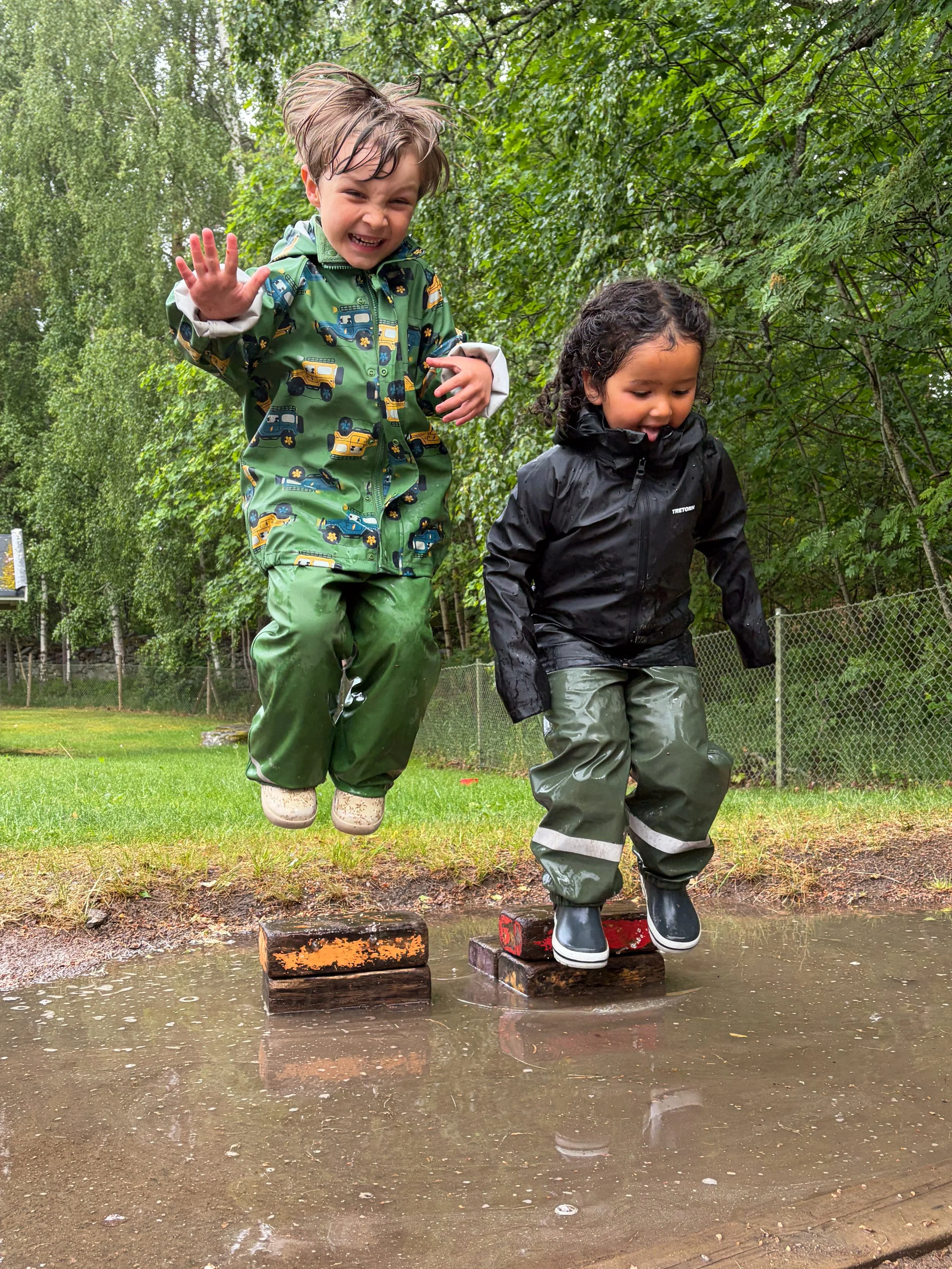 Två barn hoppar på träblockar över vattenpöl i en skog, klädda för regnväder.