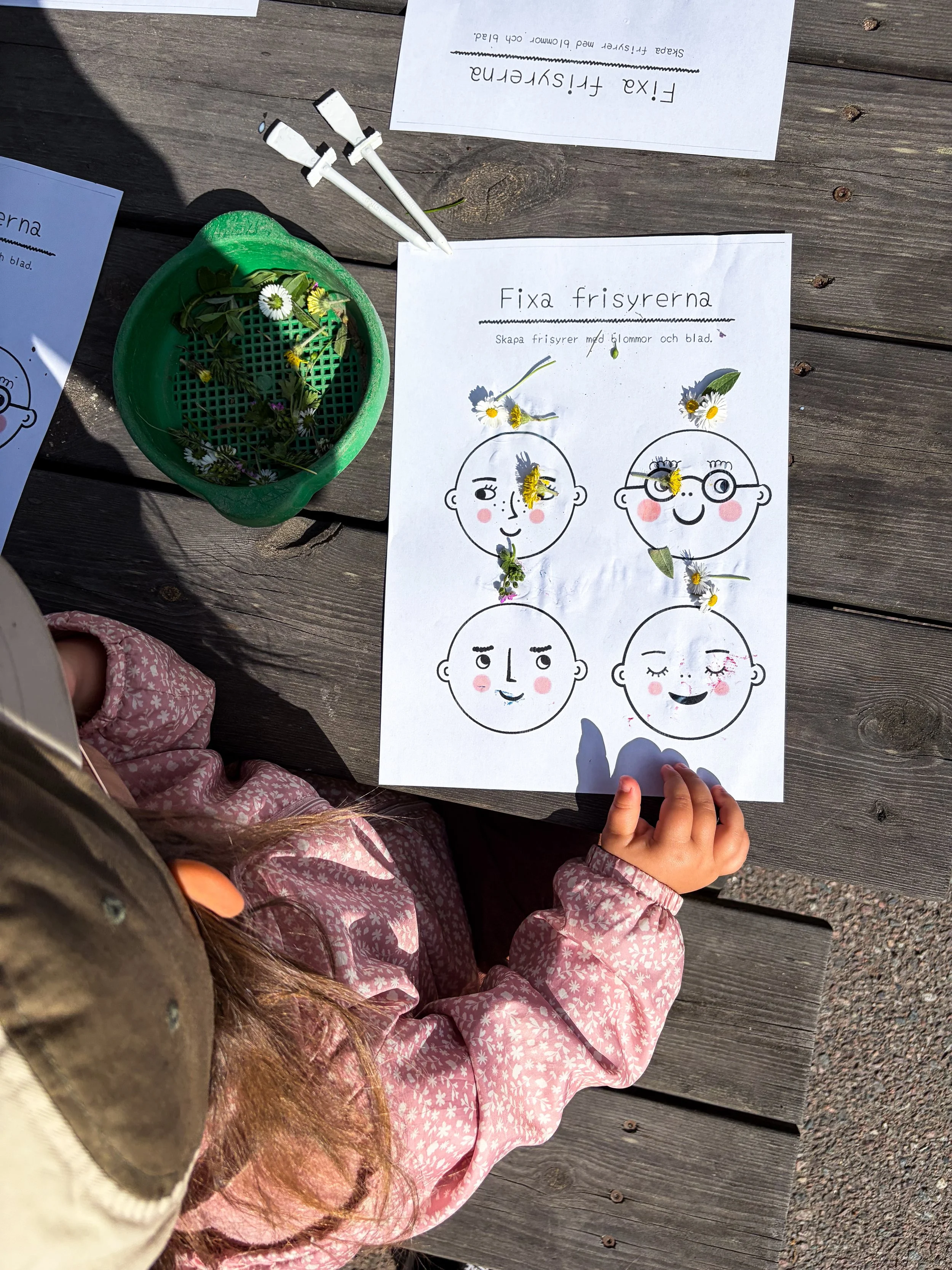 Barn i rosa jacka står vid ett bord med ett utskrivet instruktion för att skapa frisyrer med blommor och blad, samt en plastkorg fylld med olika blomblad och blad.