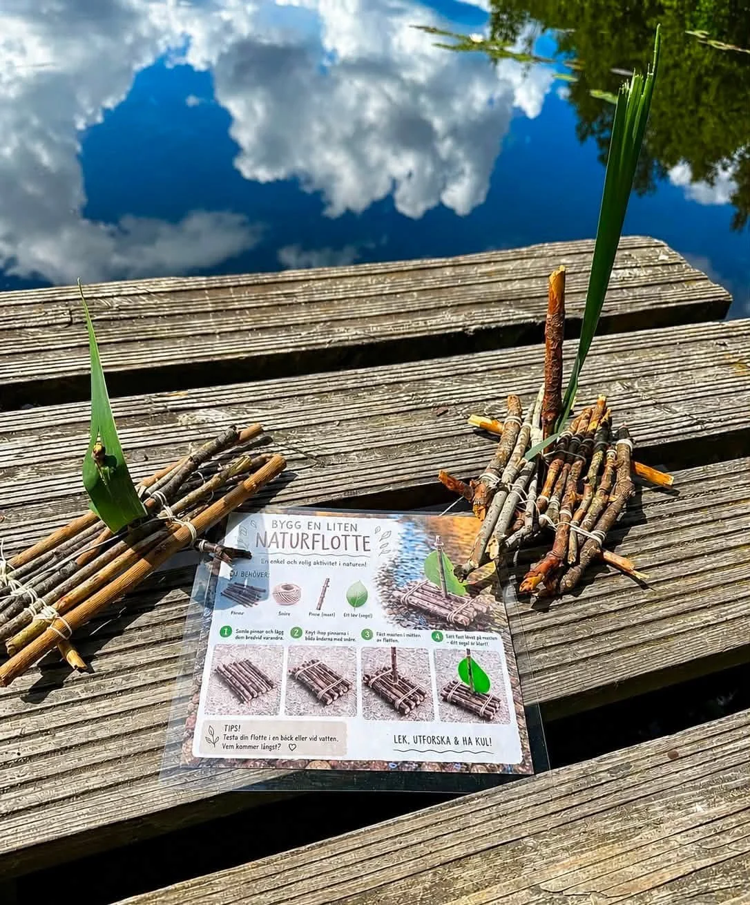 Ett informationsblad om att bygga en liten naturflotte med pinnar och blad, placerad på ett träbord vid vattnet. Rester av byggda flottebitar och ett grönt blad är synliga.