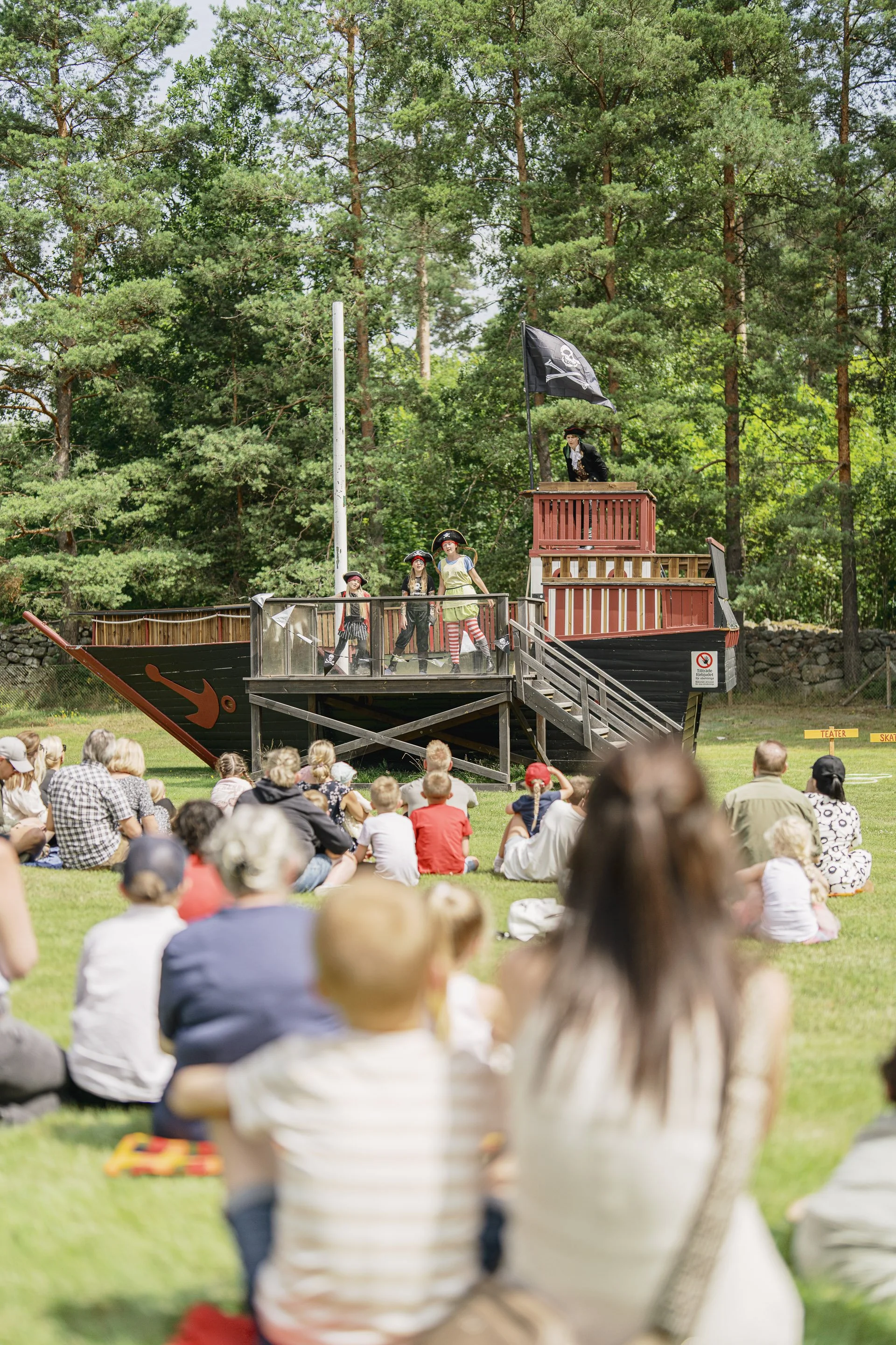 En utomhusföreställning med ett piratskepp som scen. Barn klädda som pirater står på skeppet och tittar på. En person i piratkostym står på skeppets däck. En publik av vuxna och barn sitter på gräset framför scenen, i en park med mycket grönt träd i bakgrunden.