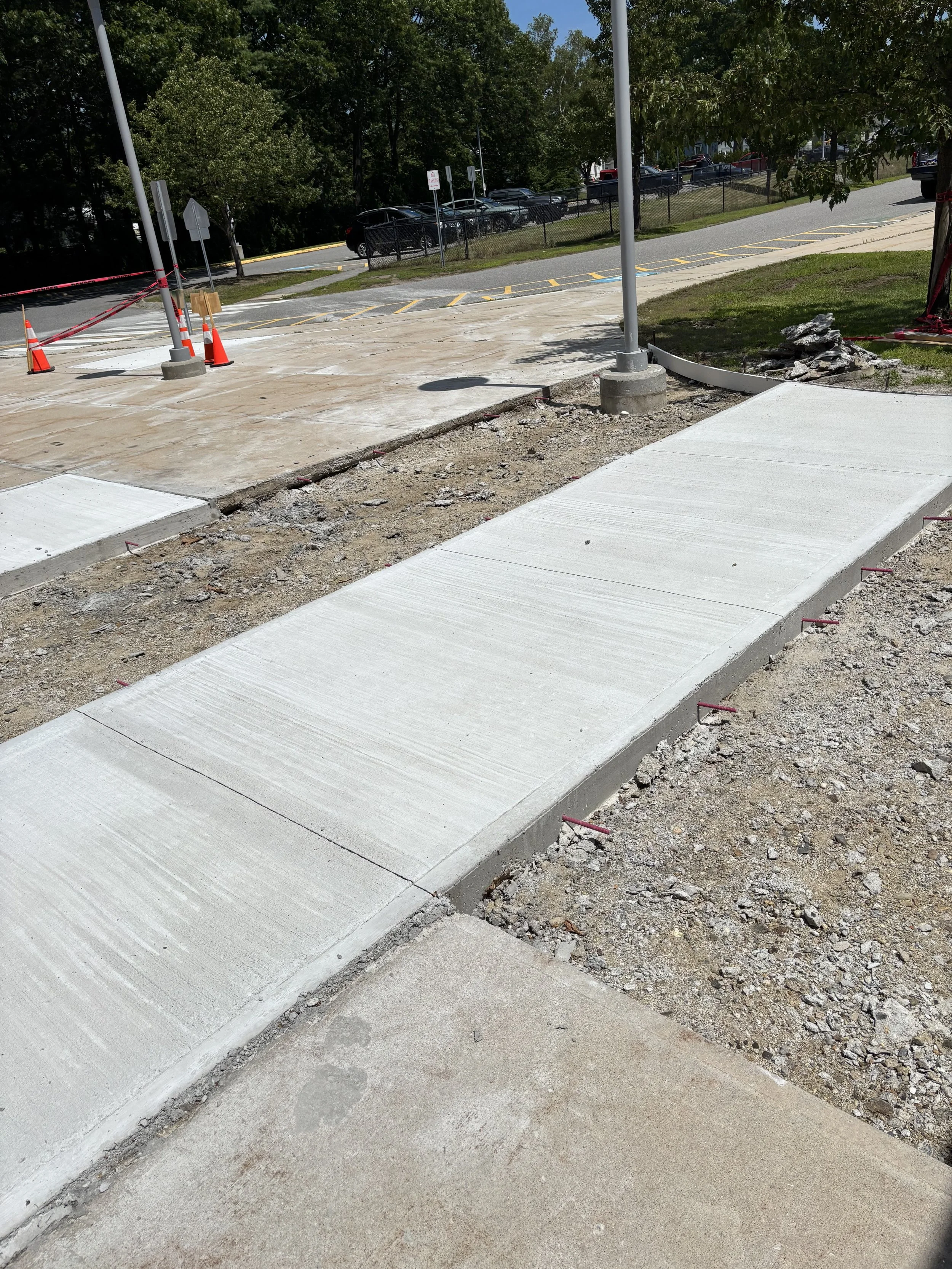Newly constructed wheelchair accessible sidewalk and curb ramp with concrete sidewalk and curb, construction tools, orange safety cones, and temporary fencing near parking lot and street. basement systems maine bone dry basement turner maine 