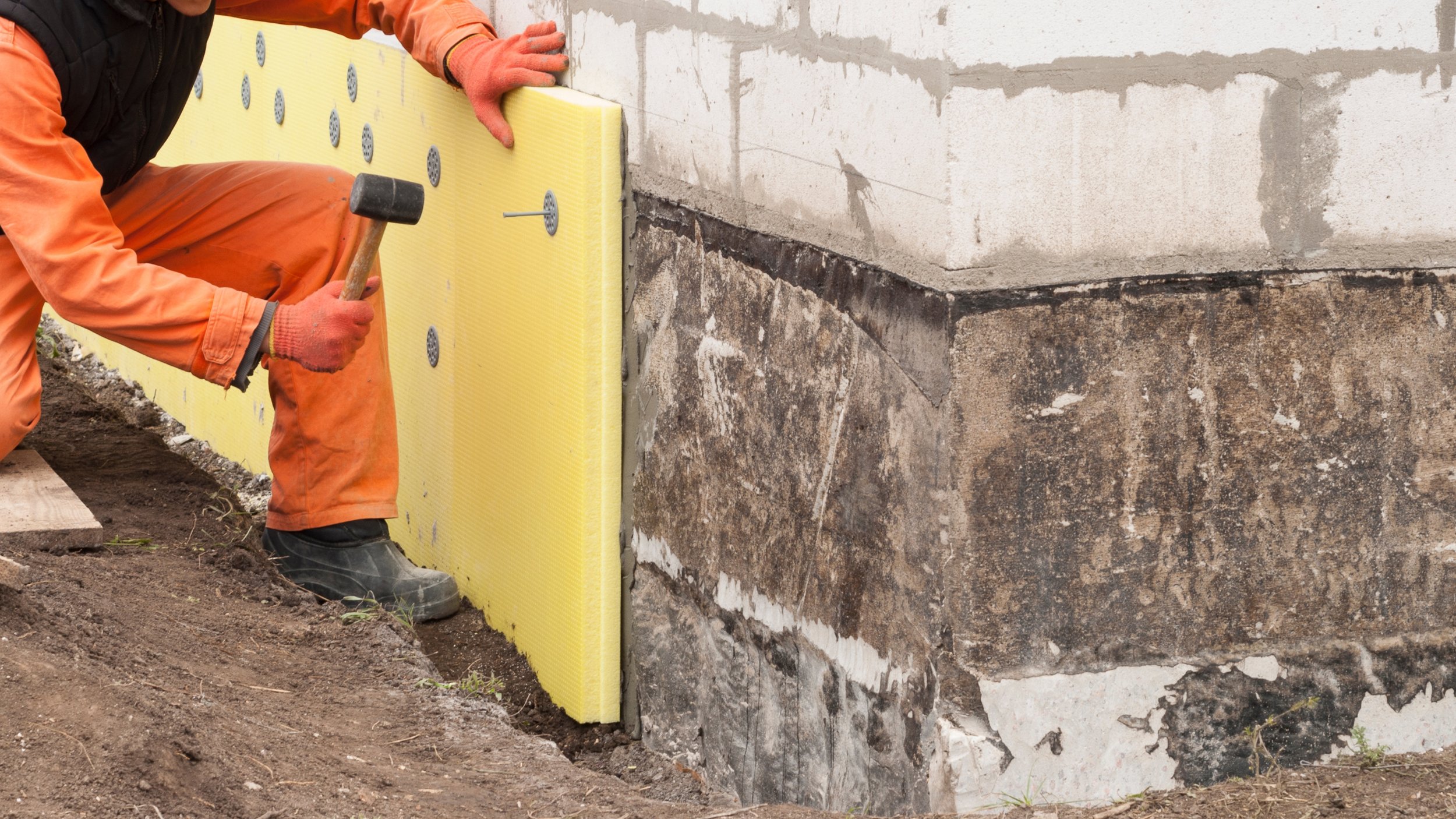A construction worker wearing orange pants, black boots, and an orange jacket installing a yellow foam insulation board along the foundation of a building with a hammer. basement systems maine bone dry basement turner maine