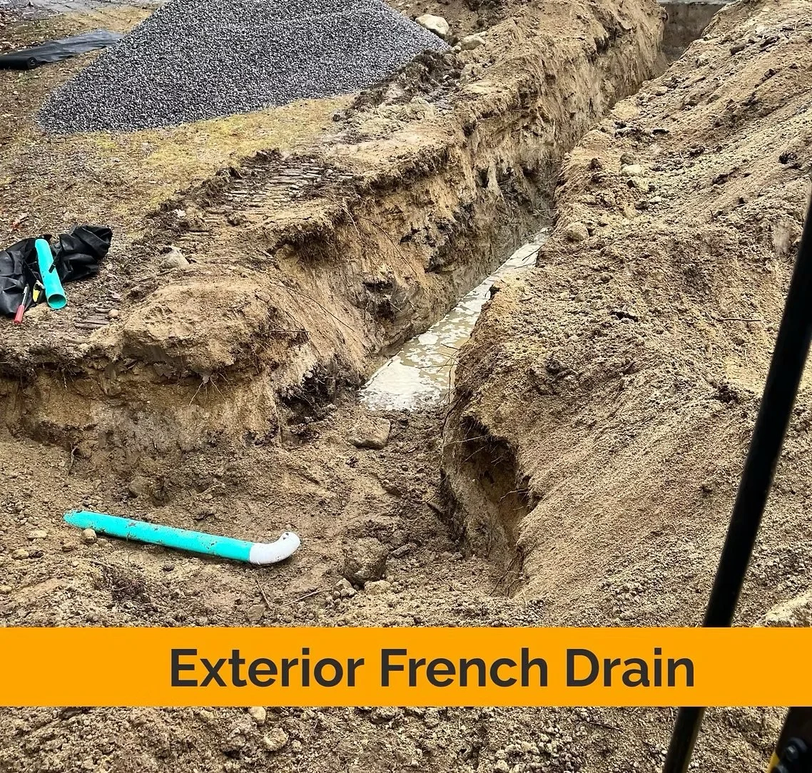A trench with a plastic pipe drainage system being installed outdoors, with tools and gravel nearby, marked as 'Exterior French Drain'. basement systems maine bone dry basement turner maine 